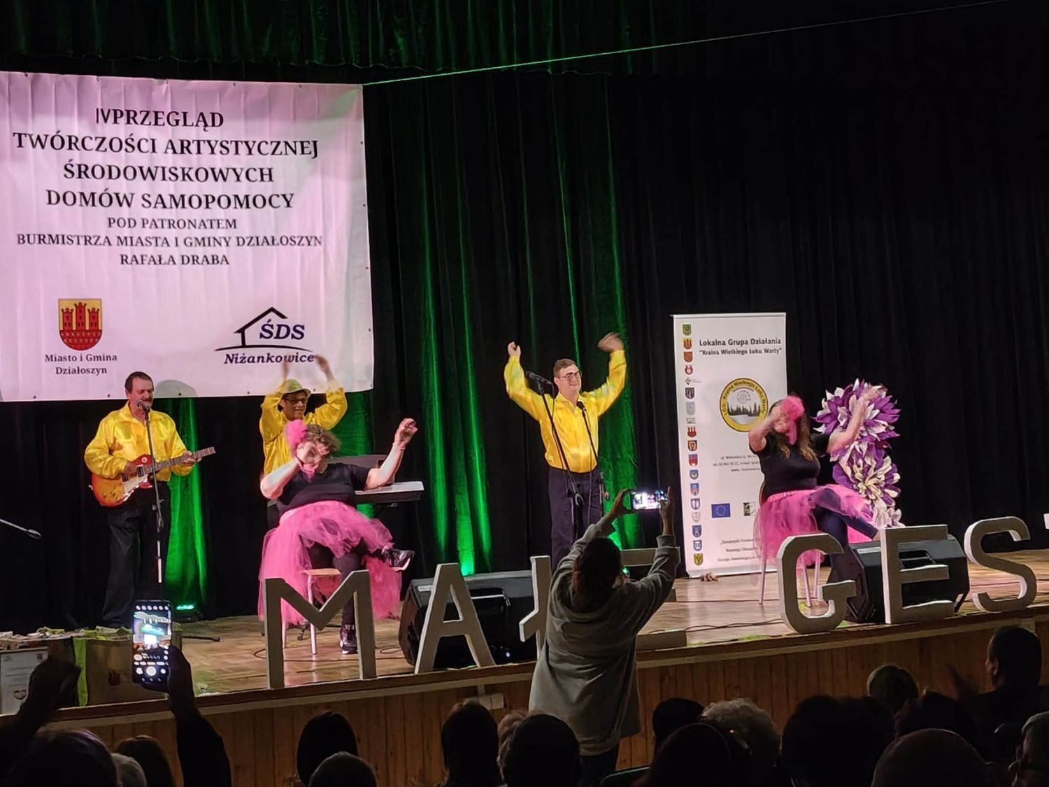 Zespół w żółtych koszulach z gitarą i tancerki w różowych spódnicach podczas występu na scenie przeglądu.