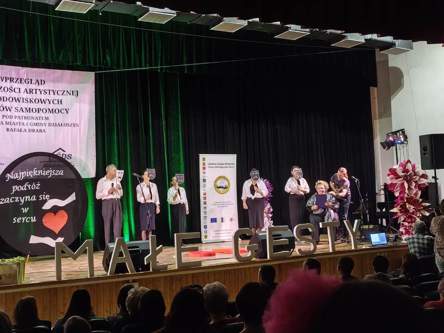 Scenka teatralna z maskami, obok hasło „Najpiękniejsza podróż zaczyna się w sercu”.
