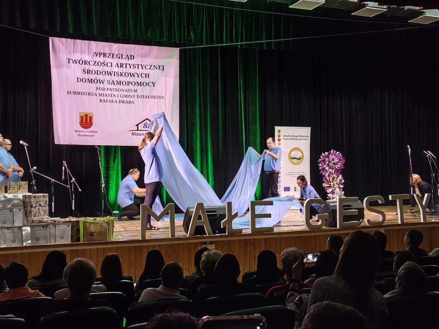 Występ z dużą błękitną tkaniną rozpościeraną przez artystów na scenie.