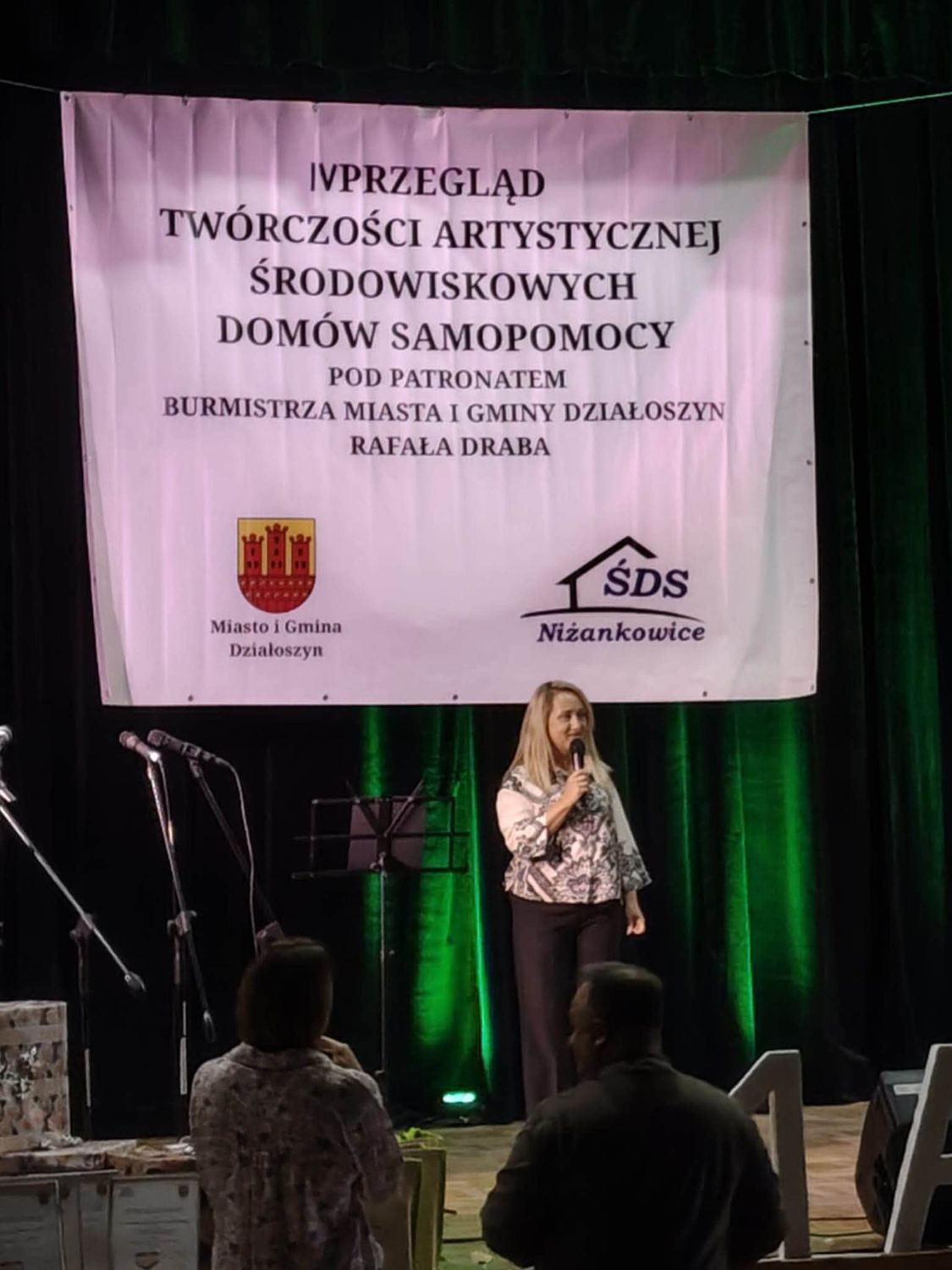 Prowadząca z mikrofonem pod banerem IV Przeglądu Twórczości Artystycznej.